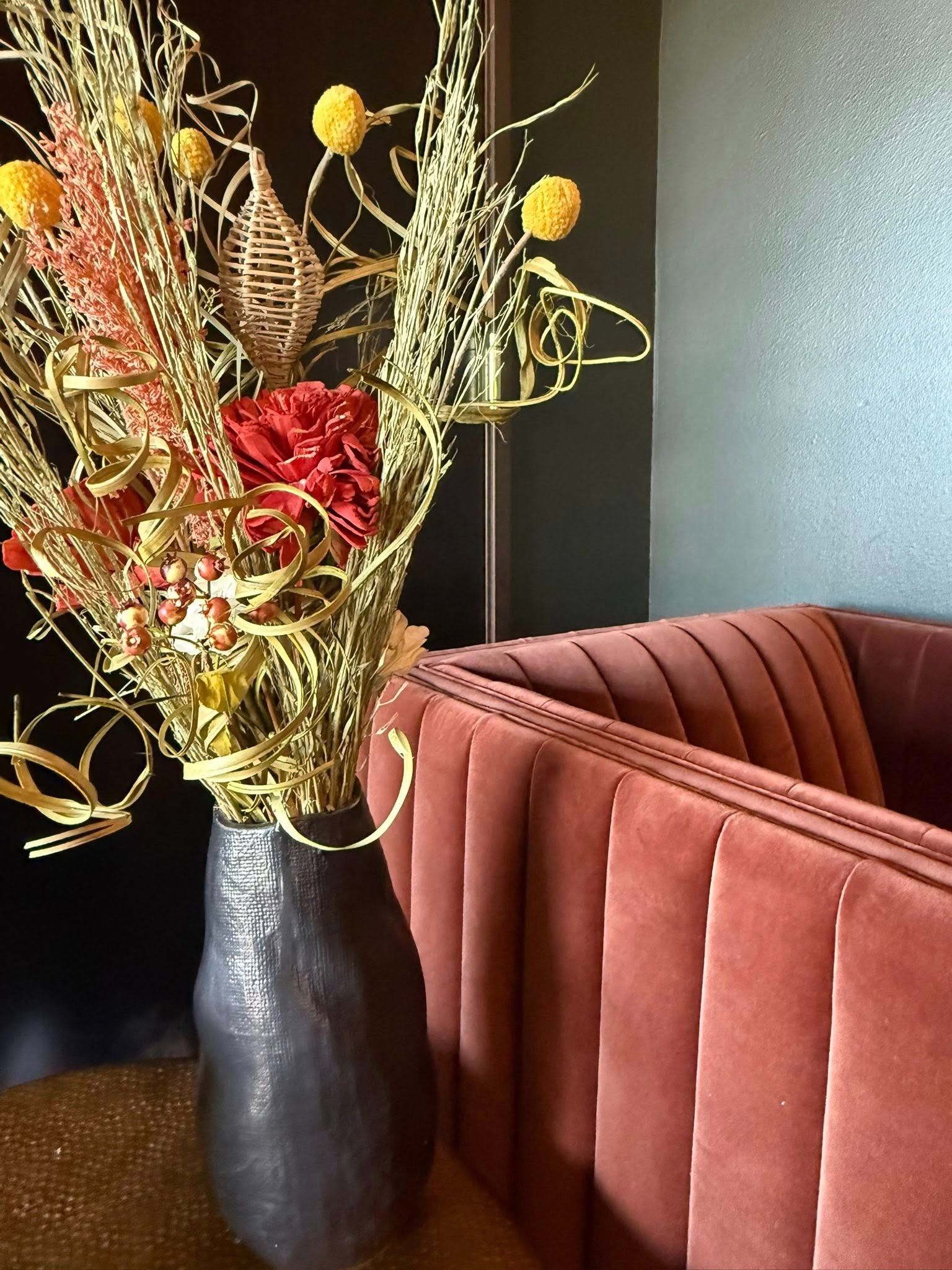 Tall black vase with dried flowers beside a rust-colored velvet sofa against a dark wall