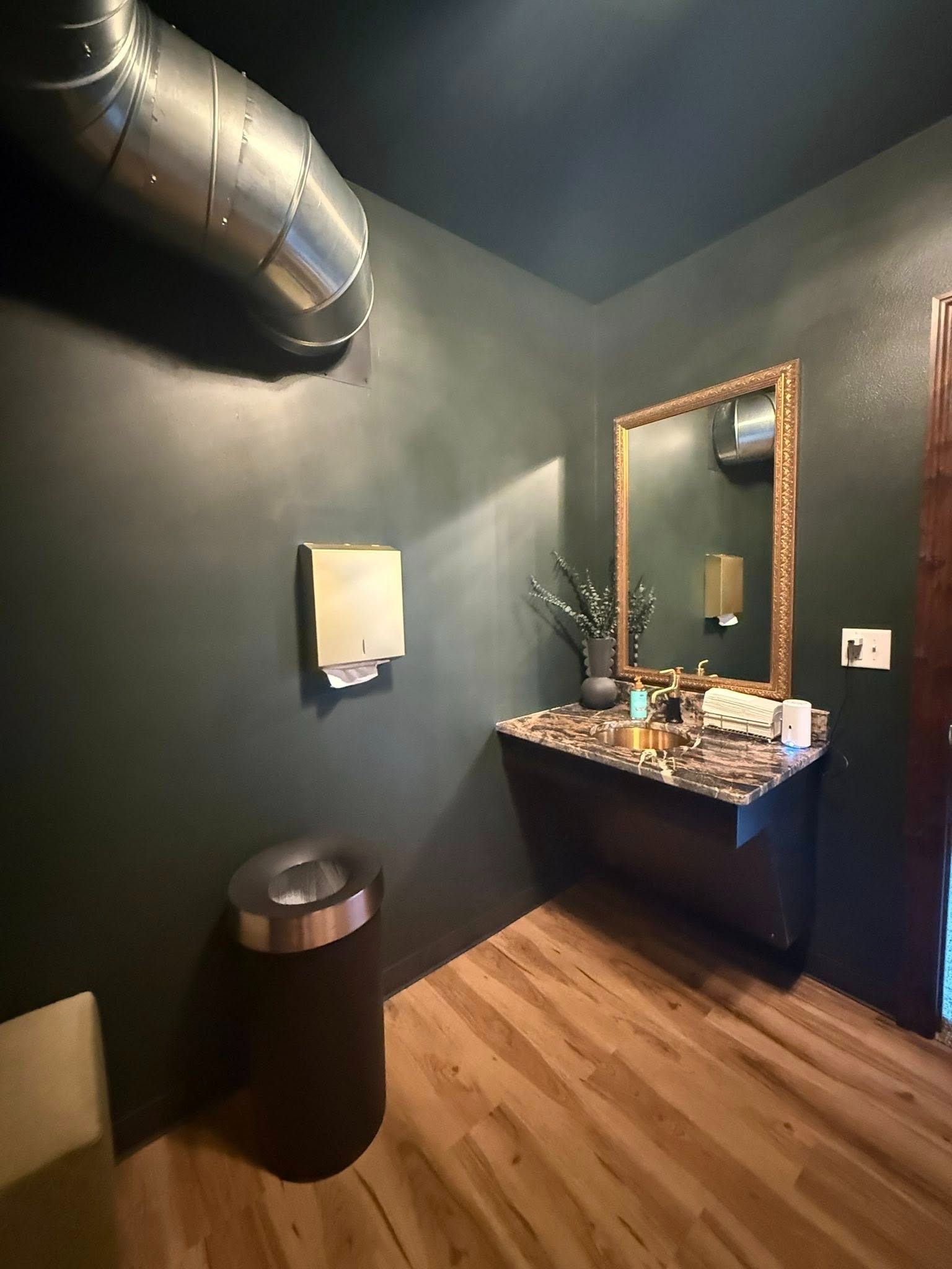 Modern bathroom with dark walls, wood floor, granite sink vanity, mirror, trash can, and exposed ductwork.