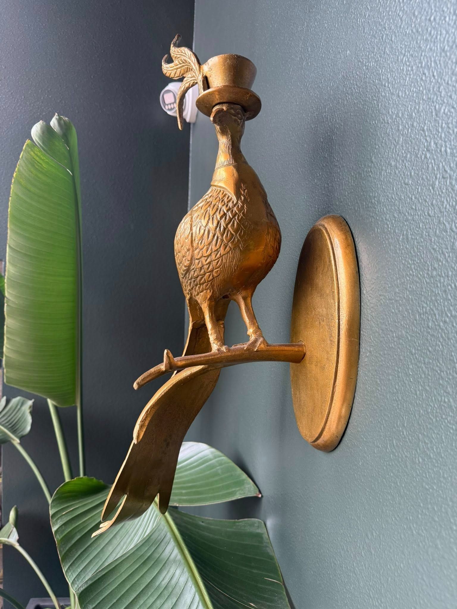 Brass bird wall sculpture on a round mount beside large green leaves.