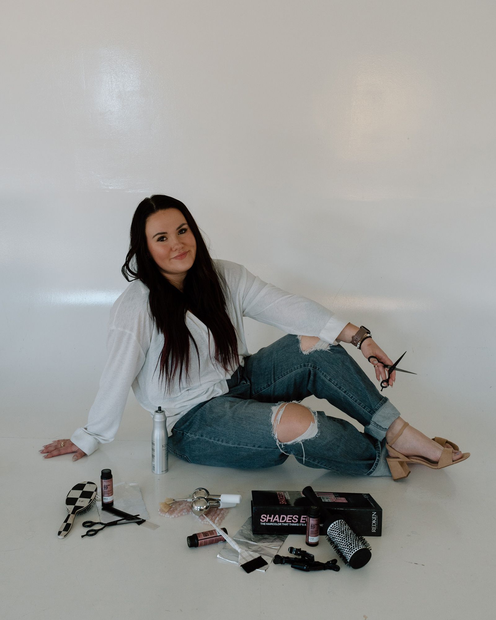 Woman seated on the floor against a white wall, surrounded by makeup products and brushes