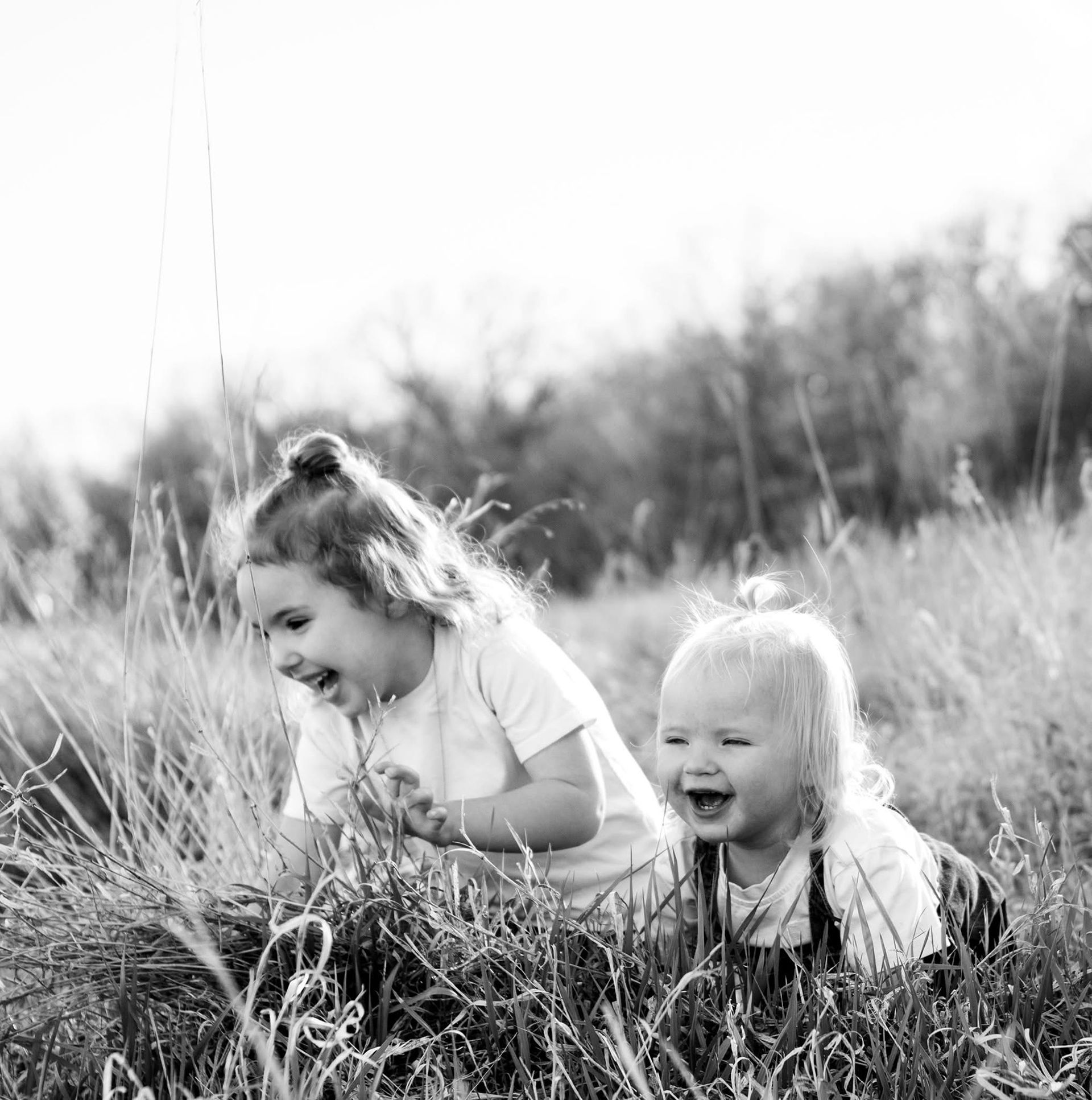 Two children lying in tall grass, smiling and laughing outdoors in black and white