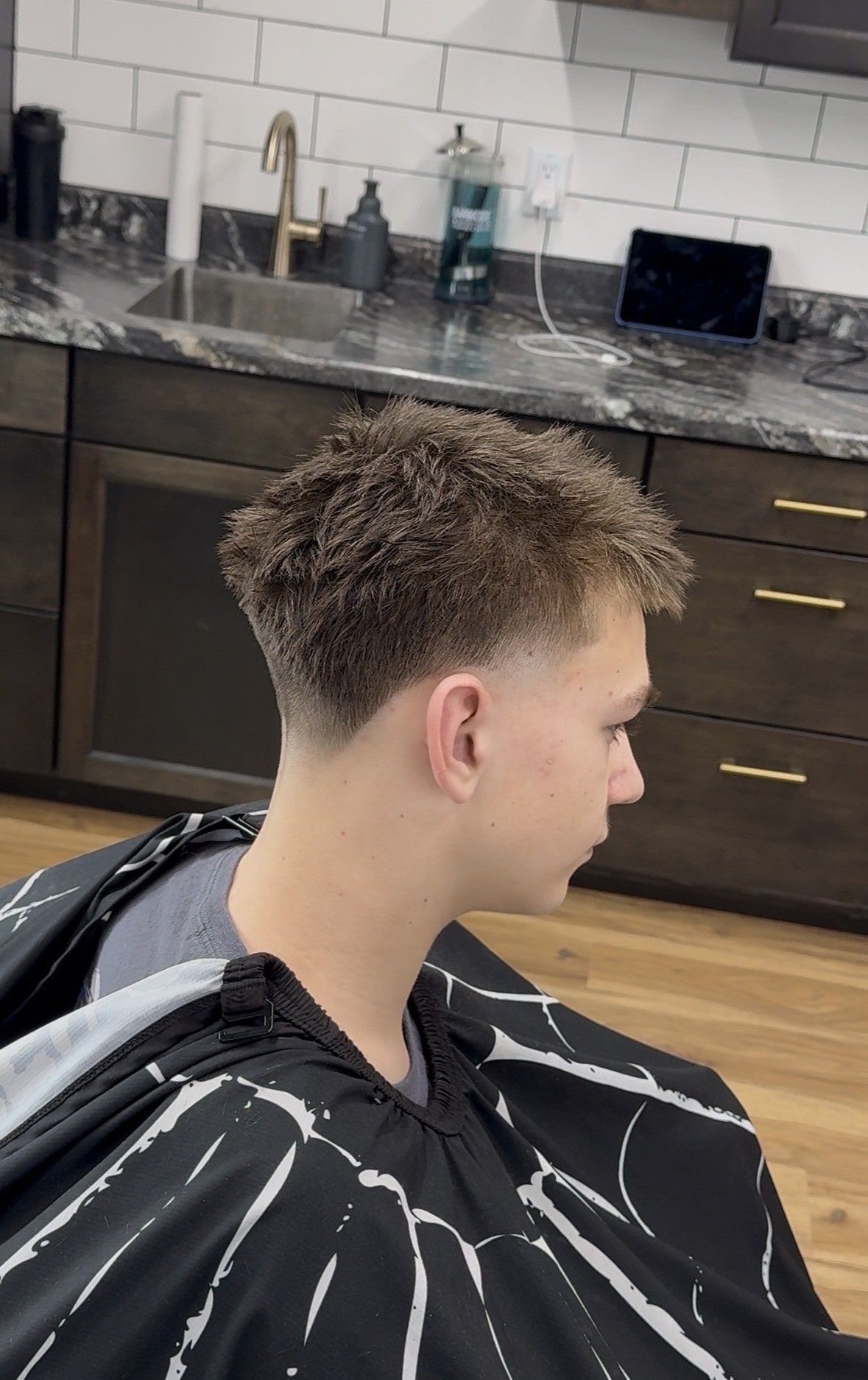 Person with short curly hair in a barber cape, seated in a kitchen-like salon setting.