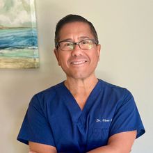 A man in a blue scrub top and glasses is smiling with his arms crossed.