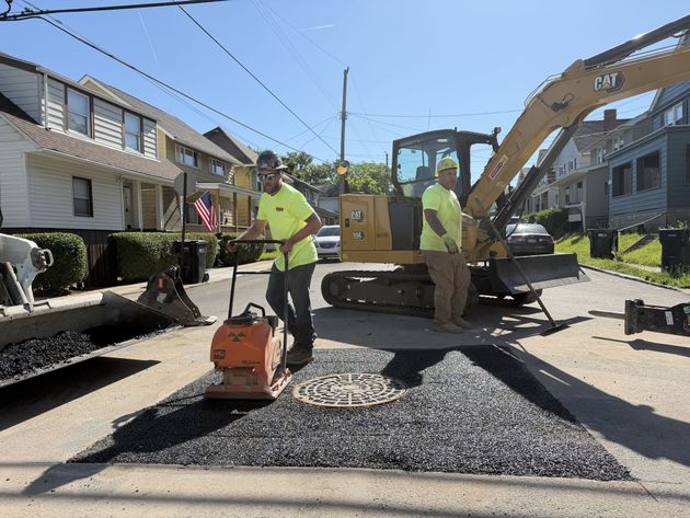 Road crew spreading gravel on a residential street with a compacting machine and excavator nearby
