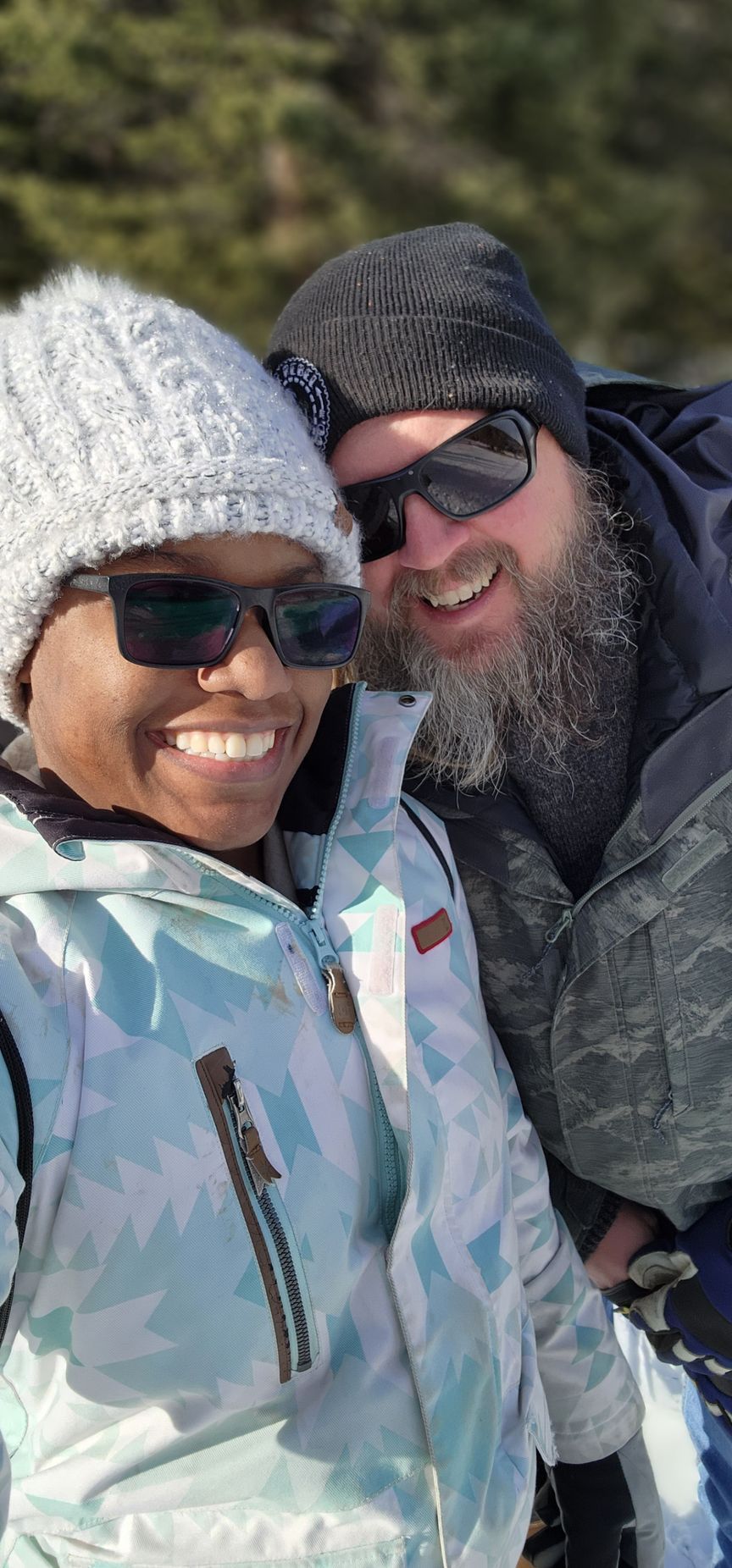 Two people wearing winter gear and sunglasses smiling together outdoors against a blurry, sunlit background.