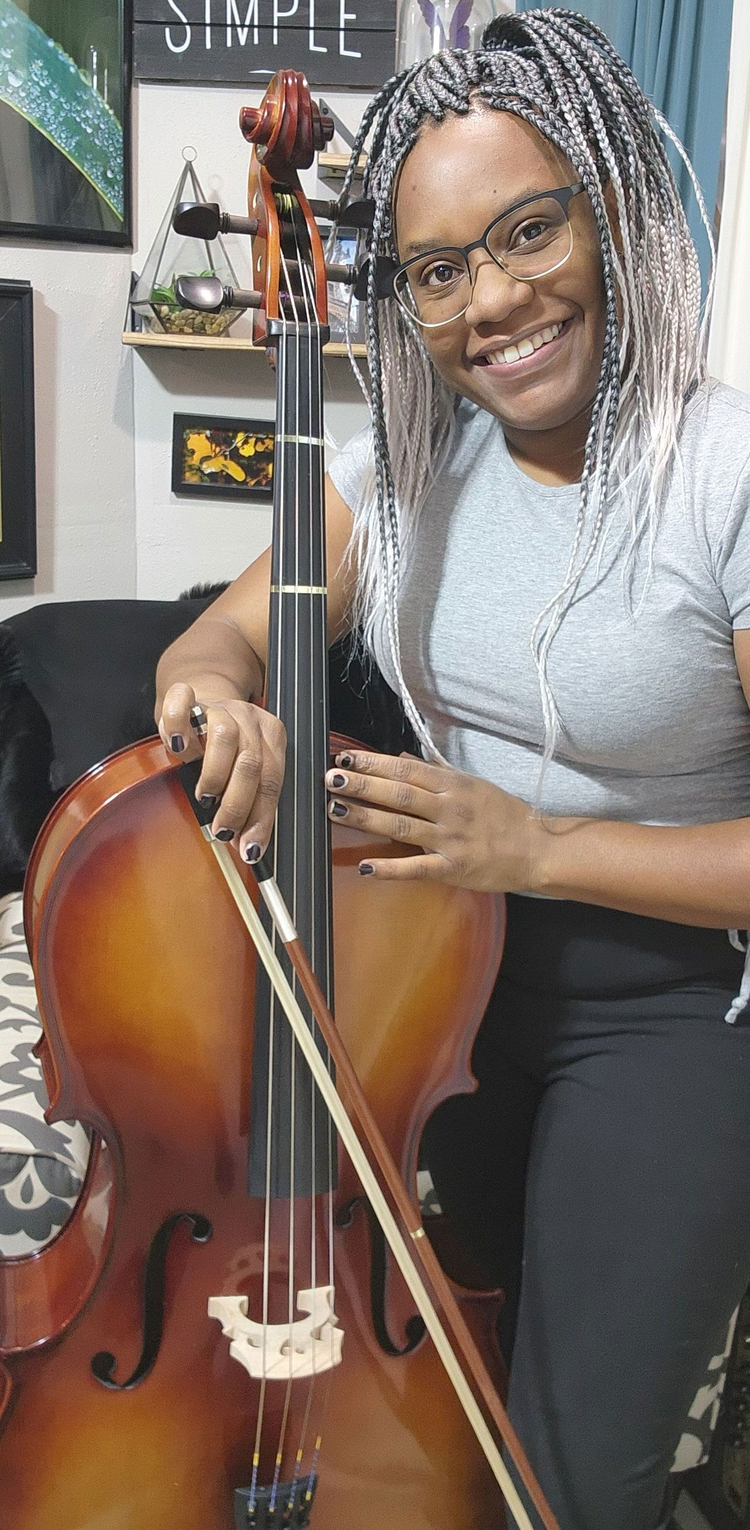 A person with braids smiles while posing with a cello and a bow in an indoor setting.