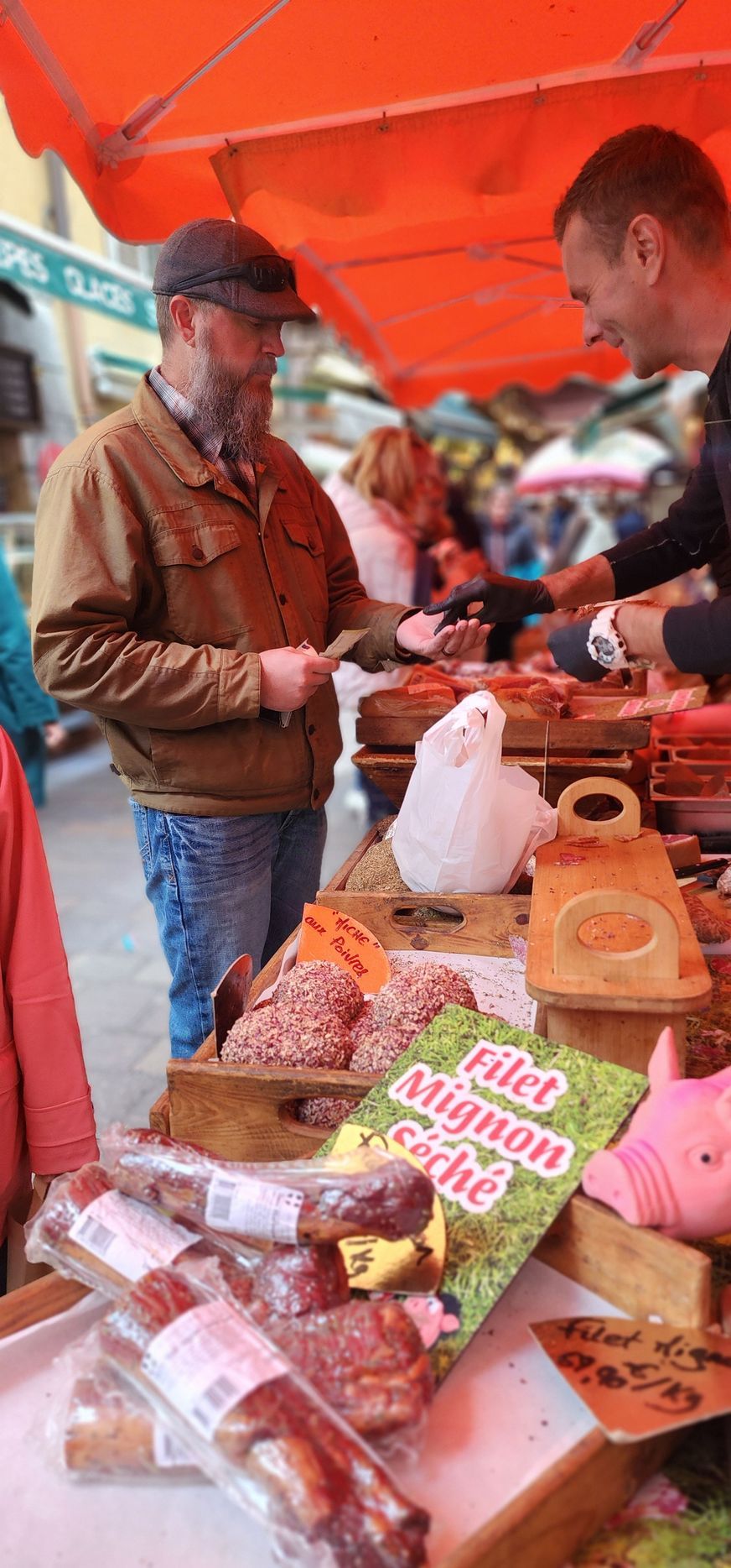 A customer interacts with a vendor at an outdoor market stall selling various meat products under a bright orange canopy.
