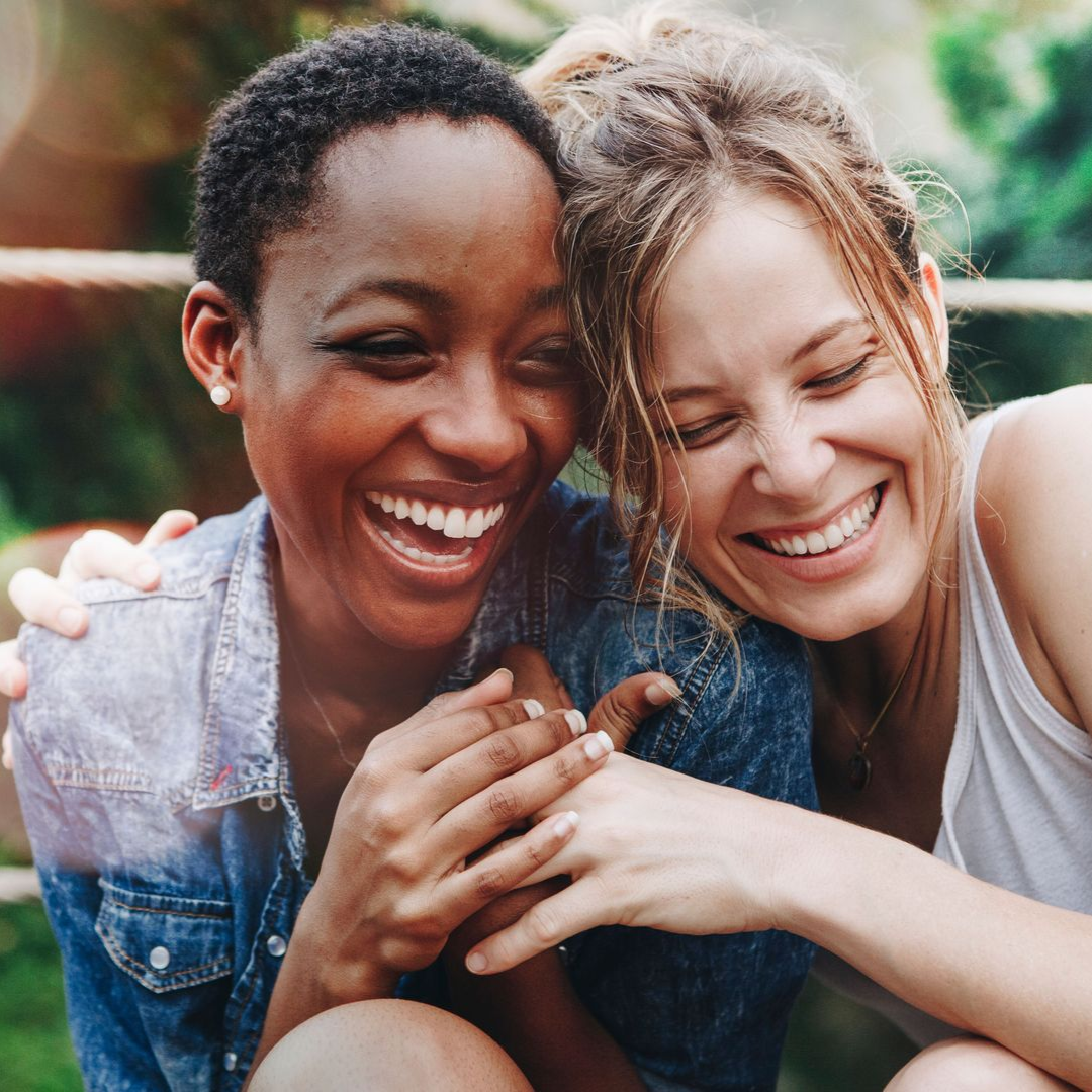 Two smiling friends hugging outdoors in a green park, with one wearing a blue top and the other a white top.