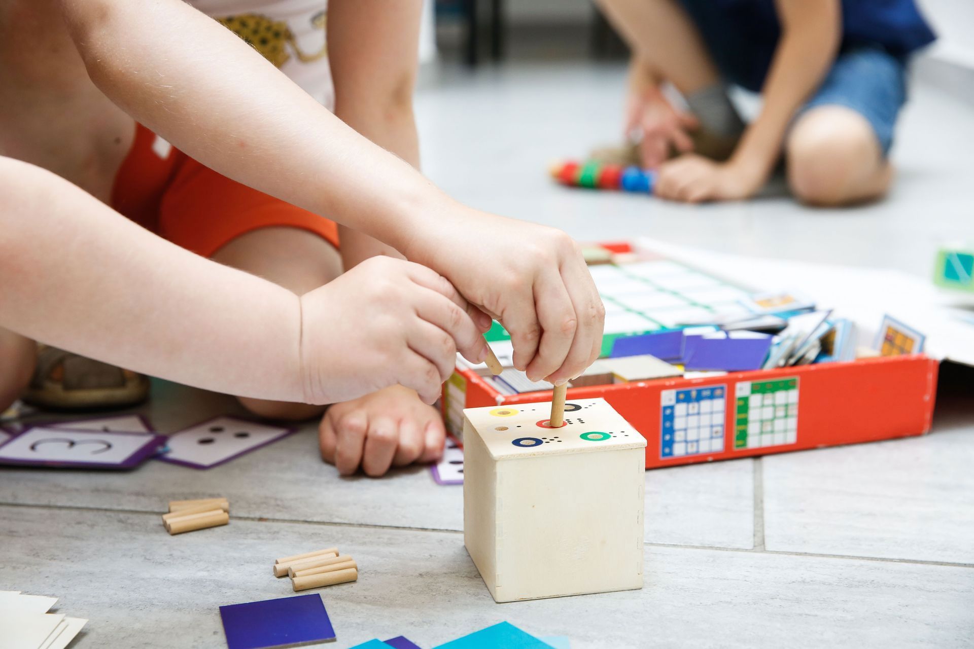 Children playing a learning game on the floor. One child inserts a peg into a wooden block.