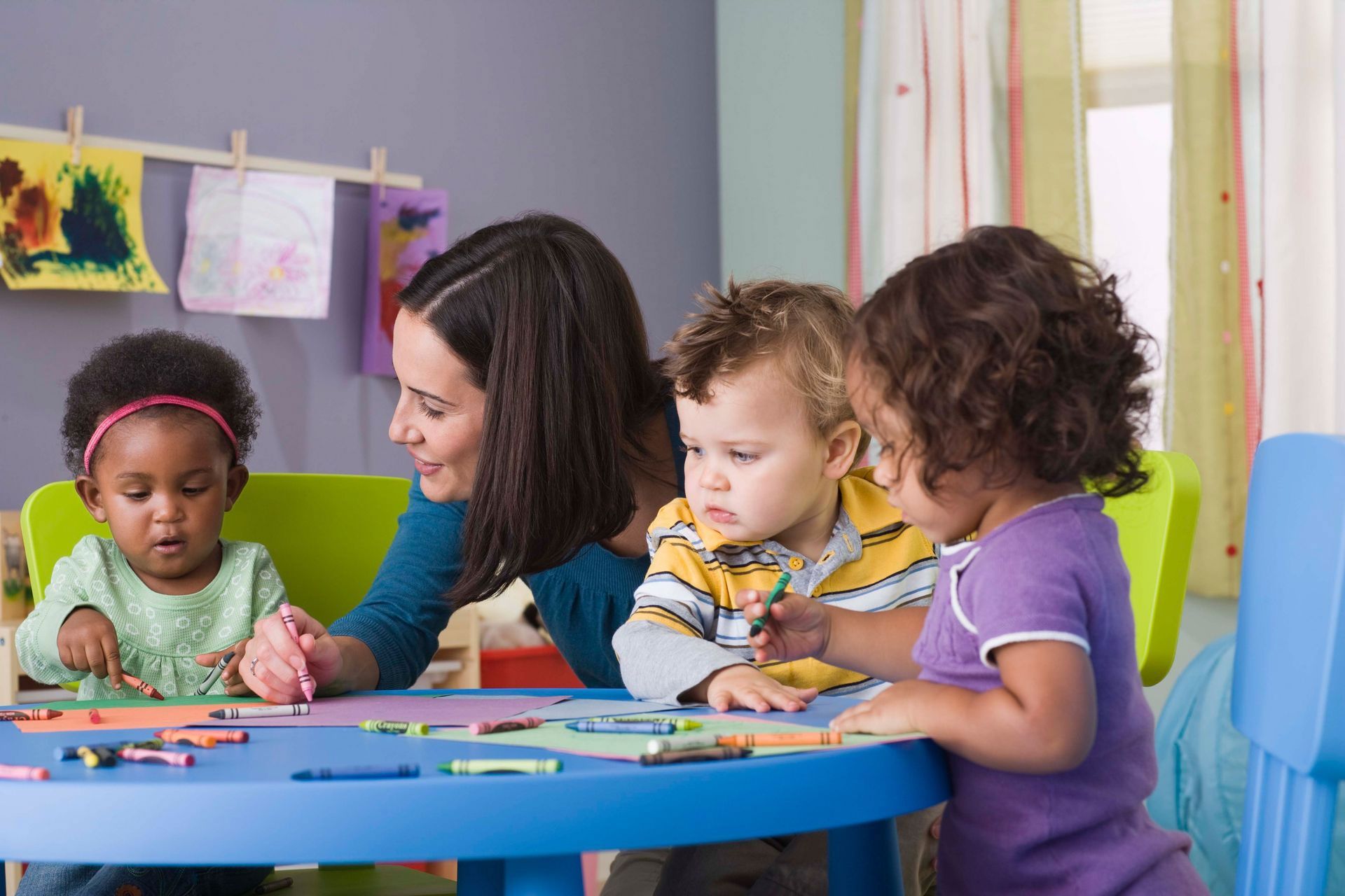 A teacher helps three toddlers with crayons at a blue table in a classroom.