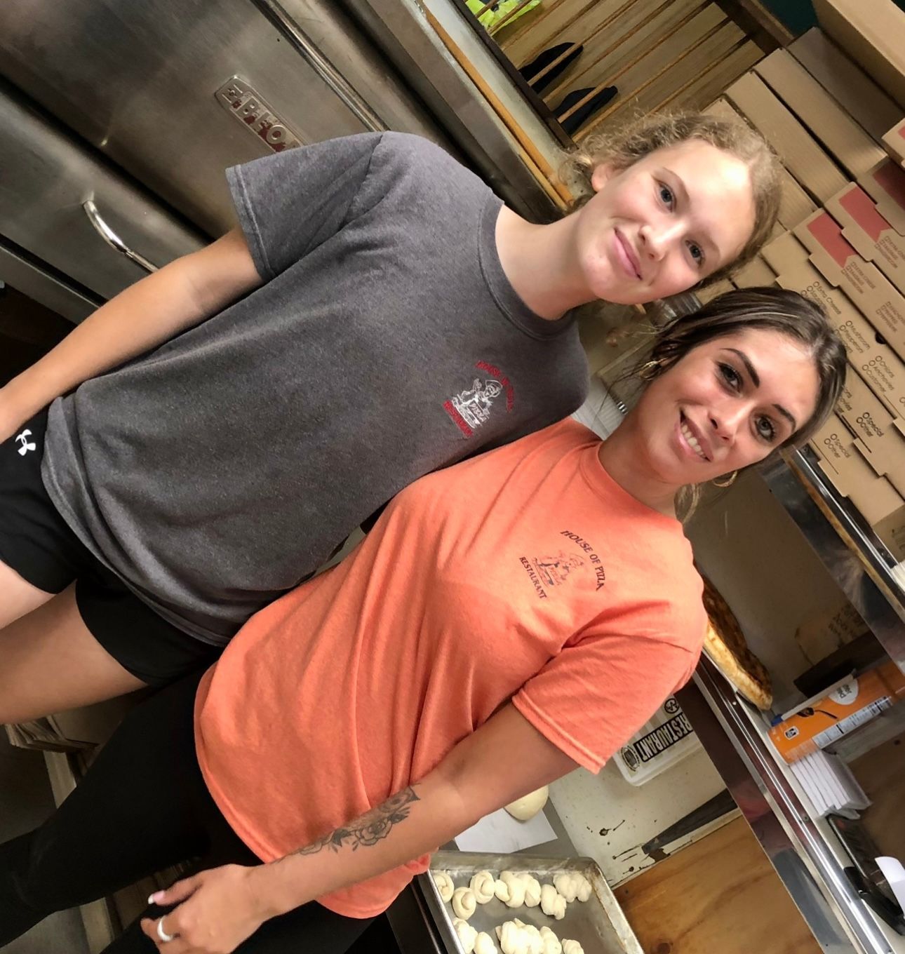 Two women standing next to each other in front of a pizza oven