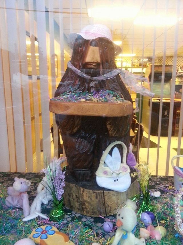 A statue of a bear wearing a pink hat is sitting on a stump surrounded by easter decorations.