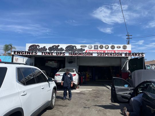 Auto repair shop exterior with parked cars and mechanics under a bright blue sky.