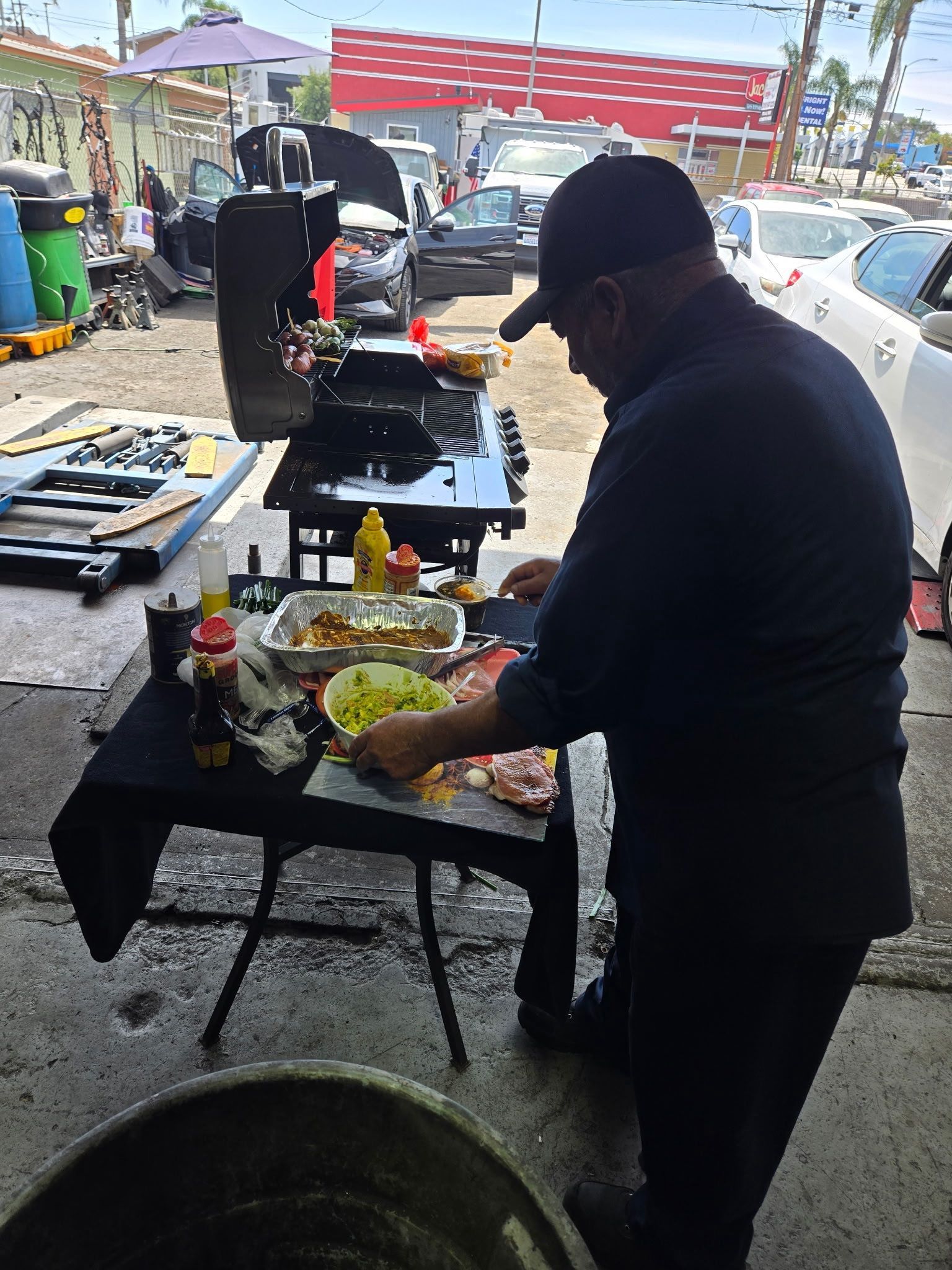Person grilling food at an outdoor market stall, with a table of ingredients and a grill in the foreground.