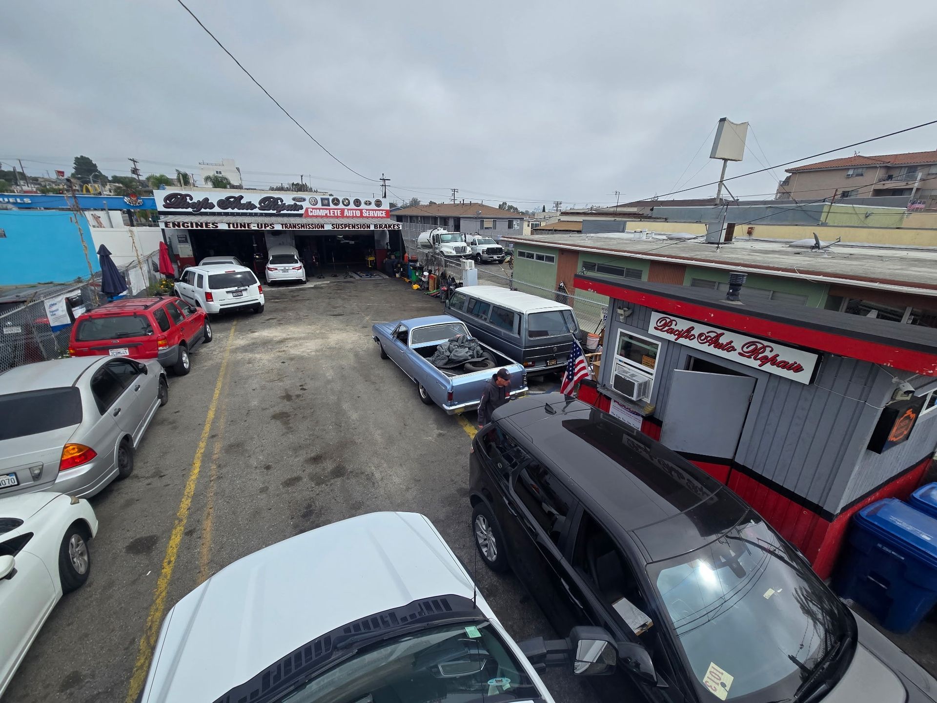 Busy car lot with parked vehicles beside small shop buildings under a cloudy sky