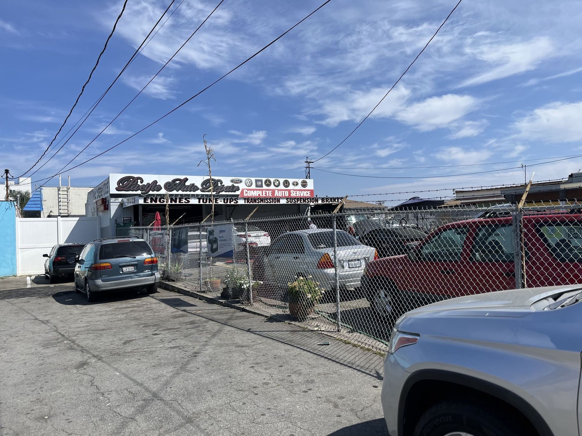 Chain-link car lot with parked vehicles, a silver truck in front, and storefronts under a blue sky.