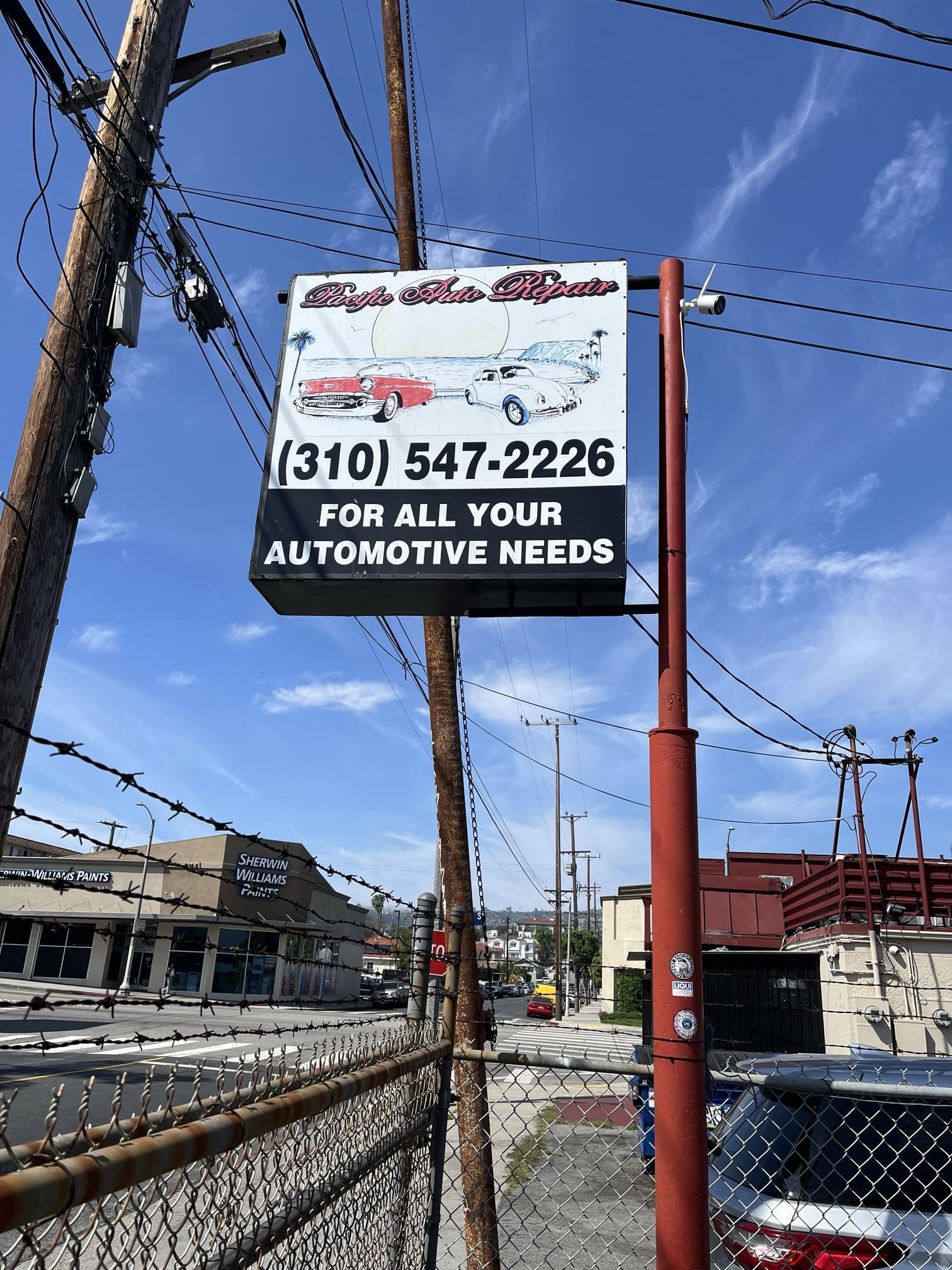 Auto repair shop sign with phone number beside a chain-link fence and power lines under a blue sky.