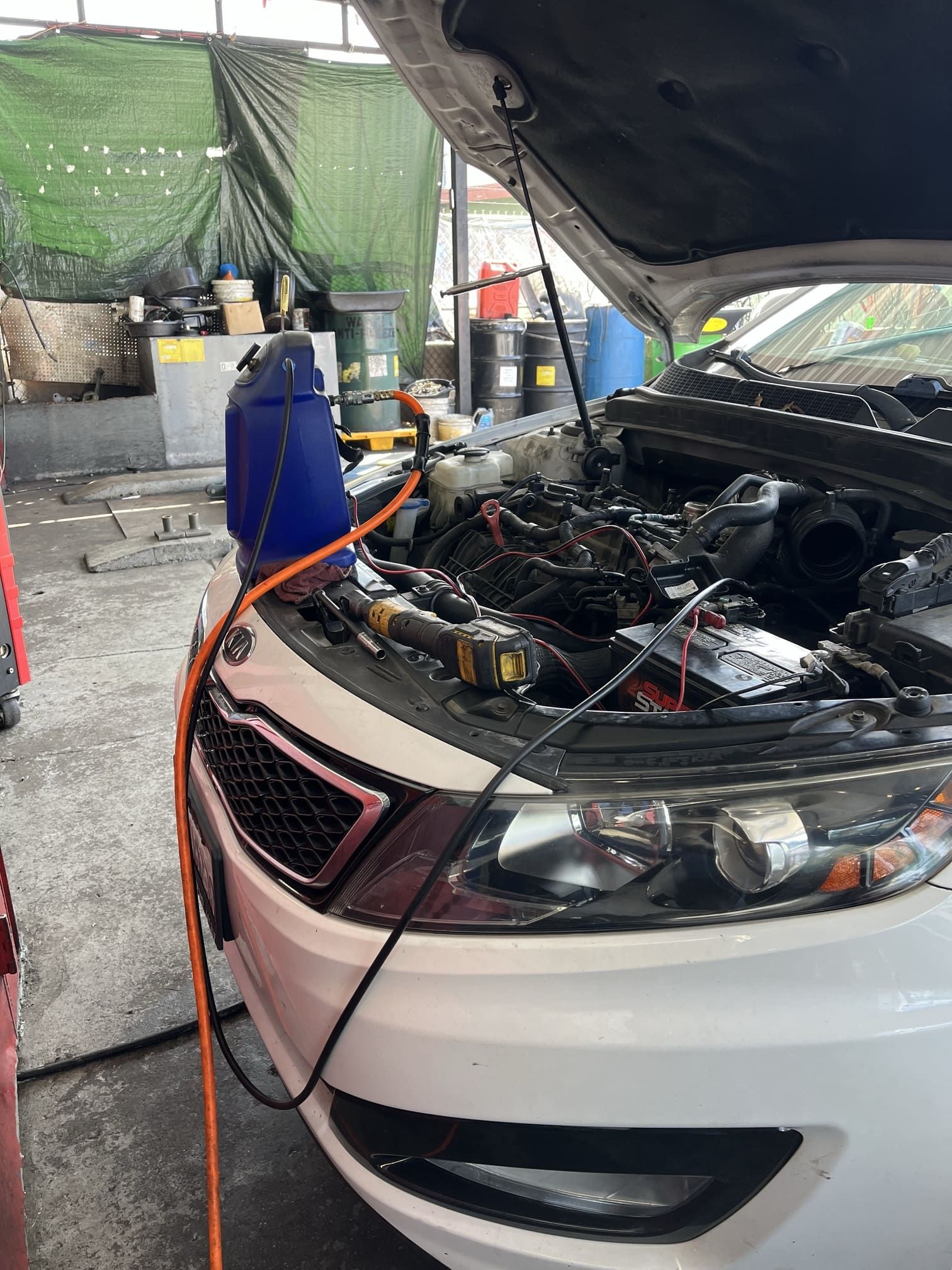 White car with hood open in a garage, showing engine repair equipment and orange hoses.