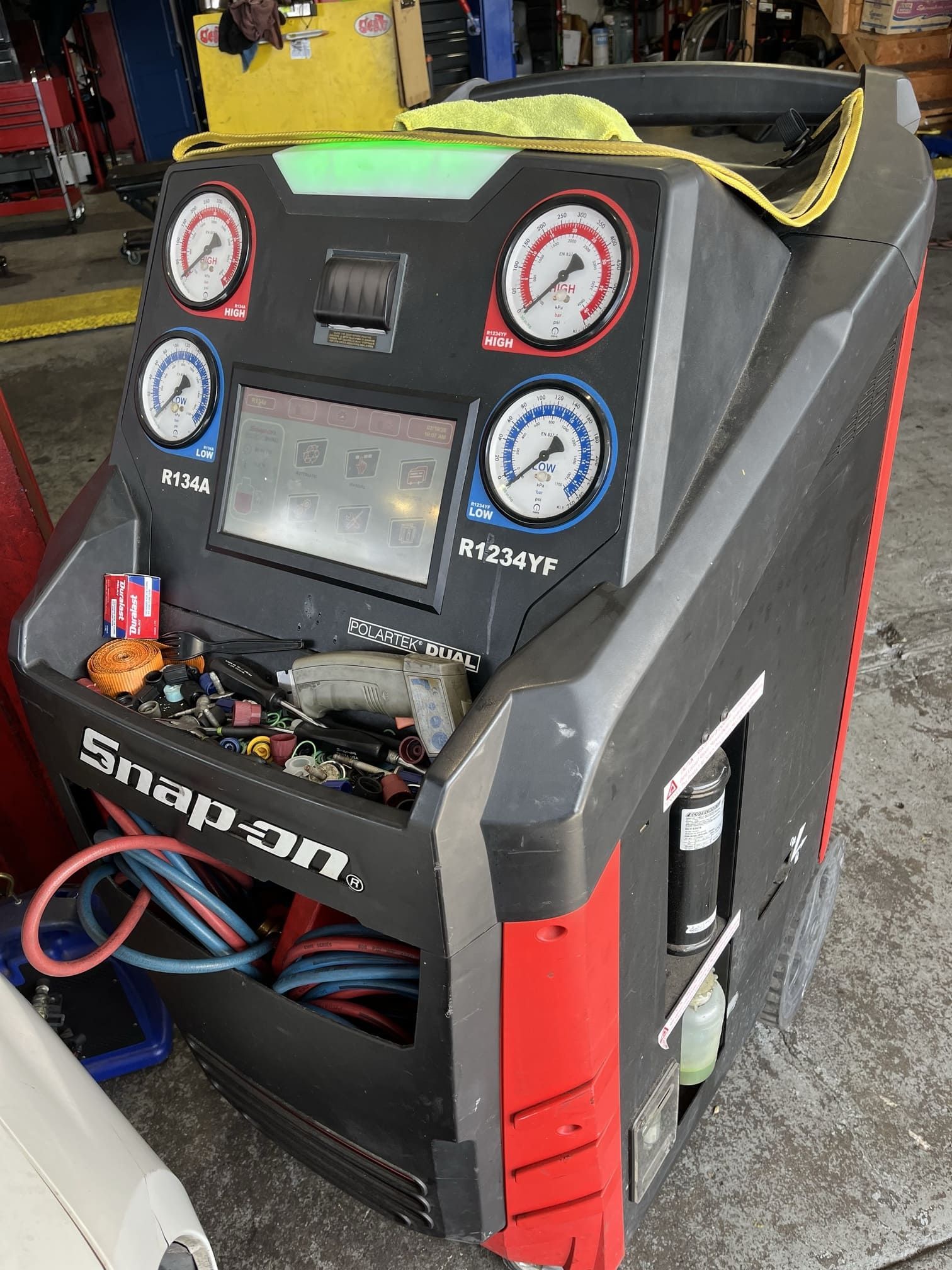 Black-and-red automotive A/C service machine with gauges, hoses, and a digital display in a workshop