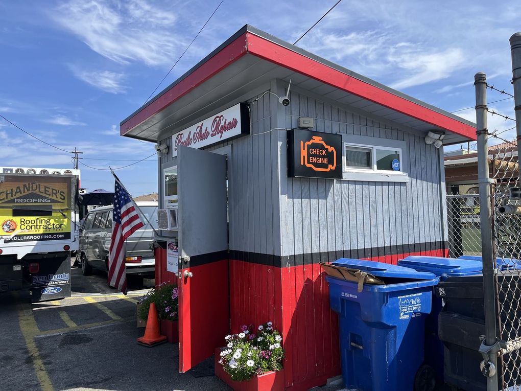Small roadside booth with red trim, neon sign, trash bins, and flowers under a sunny sky