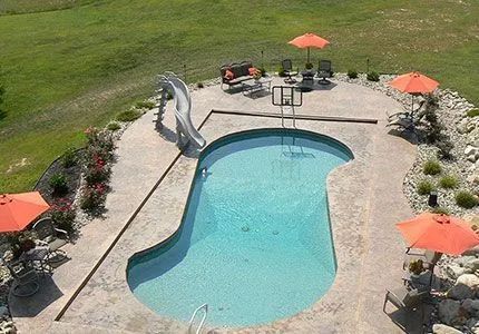 An aerial view of a kidney-shaped swimming pool with a slide, surrounded by a concrete patio, umbrellas, and lawn.