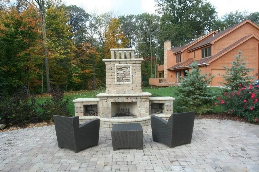 Stone fireplace with seating on a patio in front of a brick house and trees.