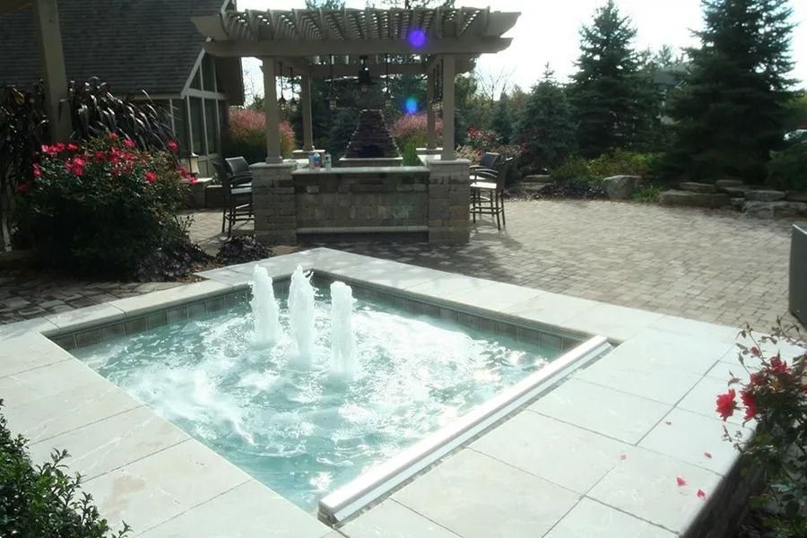 Square fountain with water jets, set in a patio with a pergola-covered outdoor kitchen.