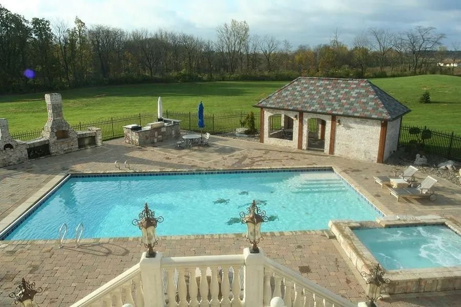 Backyard pool area with pool, hot tub, gazebo, and outdoor kitchen. Green lawn and trees in the background.