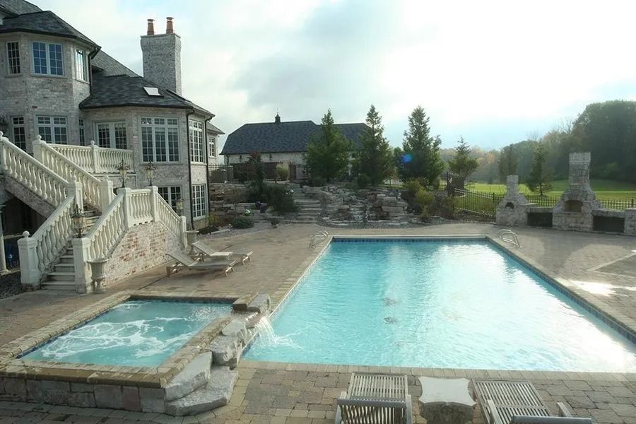 Luxurious stone house with pool, hot tub, and outdoor fireplace. Cloudy sky and manicured lawn.