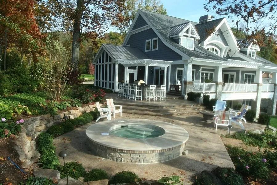 House with porch overlooking a patio with hot tub, surrounded by landscaping.