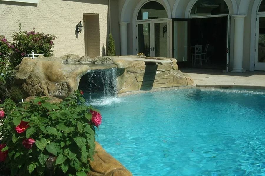 Pool with waterfall feature and a house in the background. Turquoise water, rock formation, and sunny outdoor setting.
