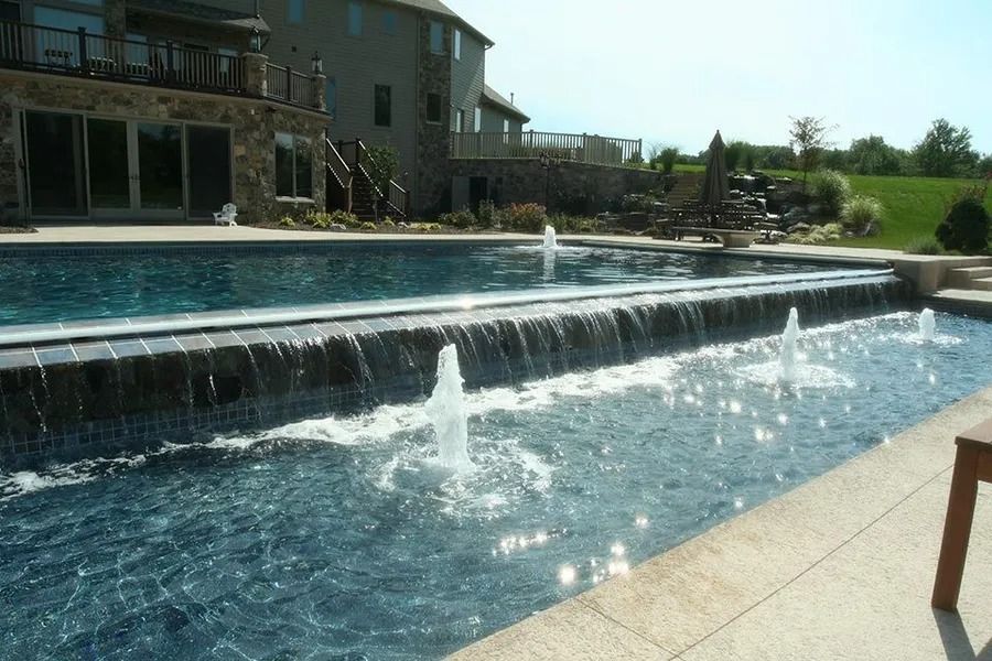 Pool with fountains, cascading waterfall, and large house in the background on a sunny day.