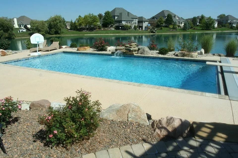 A rectangular pool with a waterfall feature overlooks a lake and houses under a clear sky.