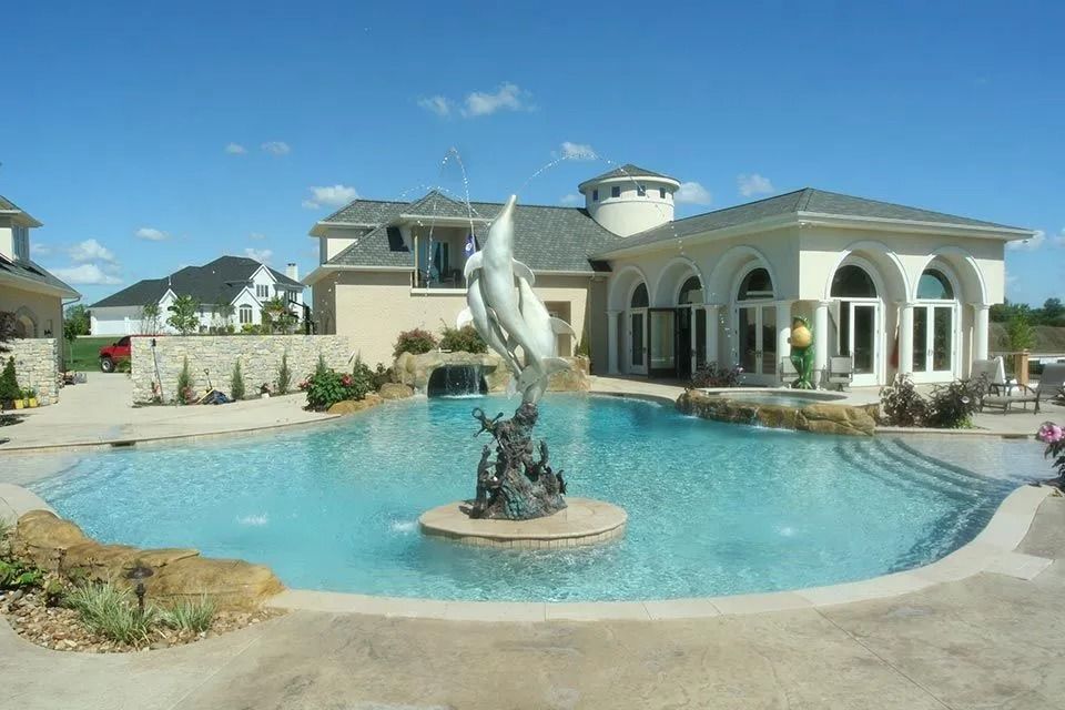 Luxurious pool with fountain, surrounded by a beige mansion and clear blue sky.