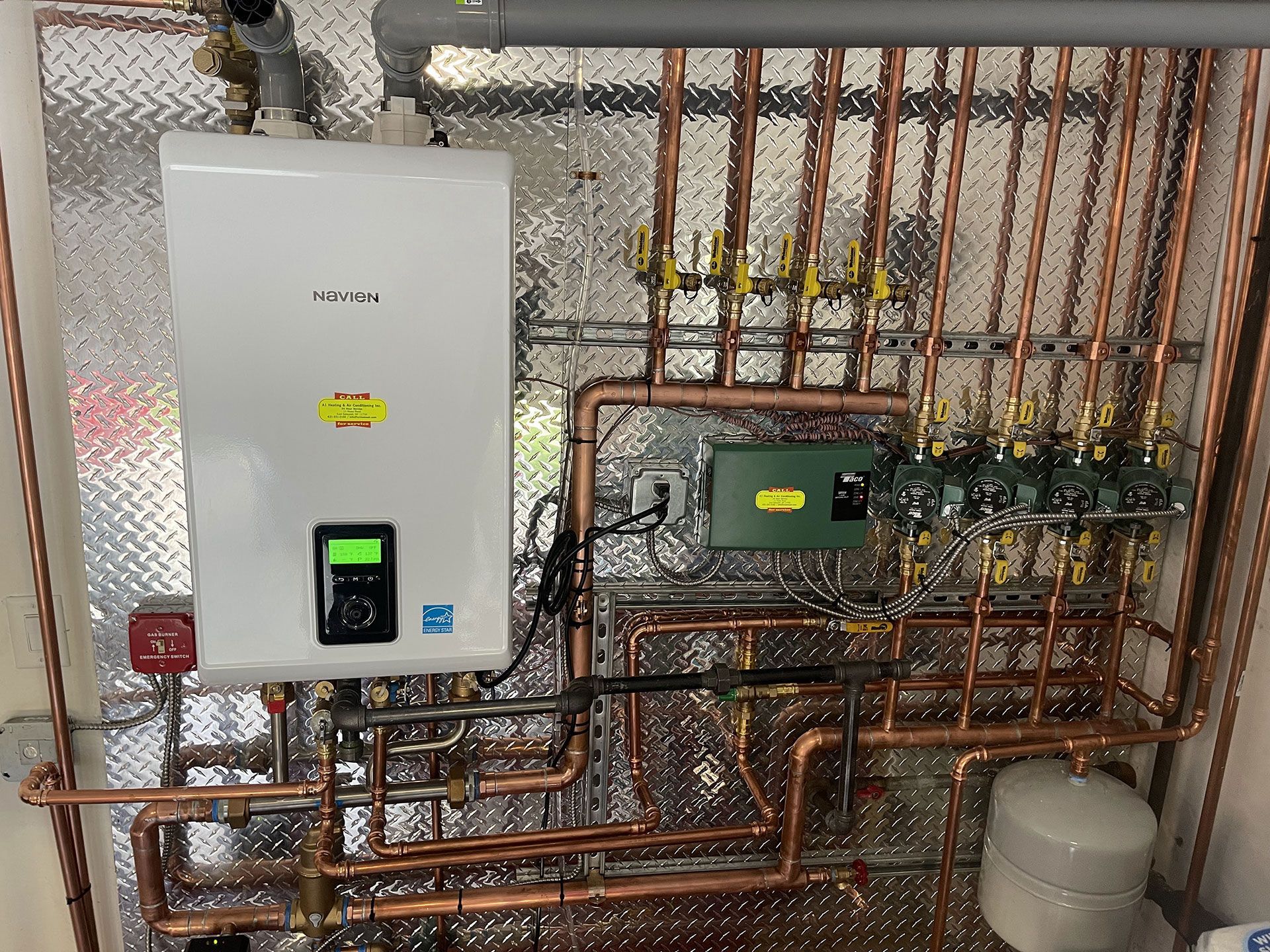 A boiler is sitting on top of a wall surrounded by copper pipes.