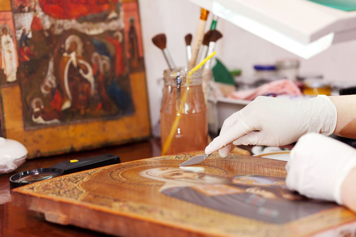 Gloved hands restoring a painting on a workbench with brushes and art supplies nearby