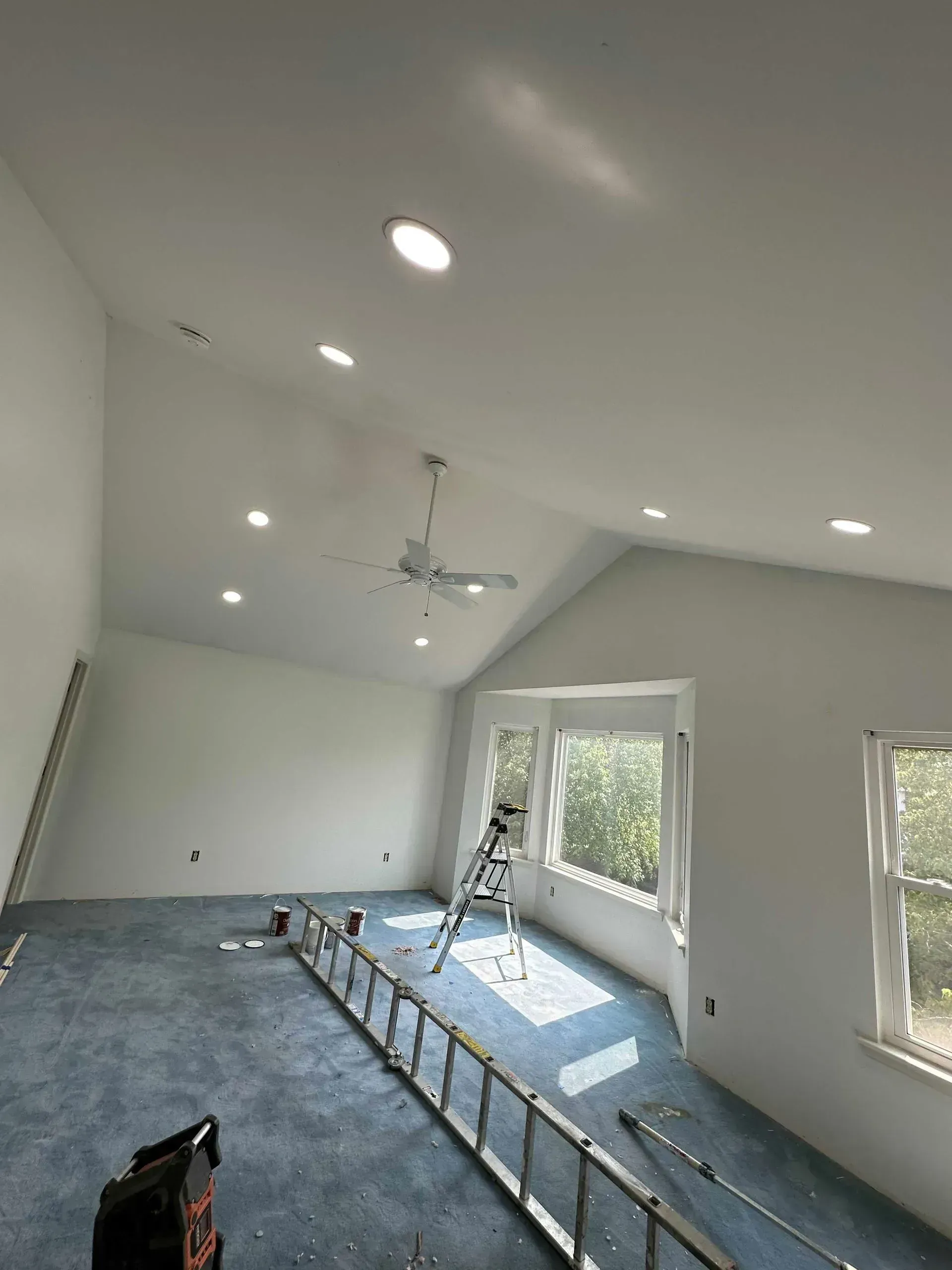 Interior of a room with a high, slanted white ceiling, recessed lights, a ceiling fan, and blue carpet