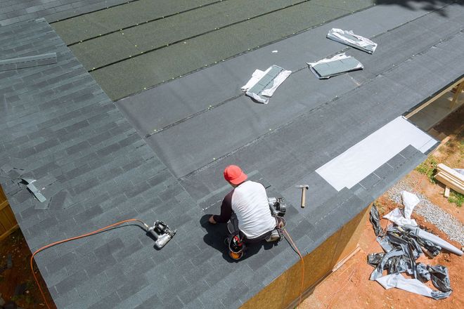 Worker installing roofing on a sloped roof with gray shingles and scattered materials.