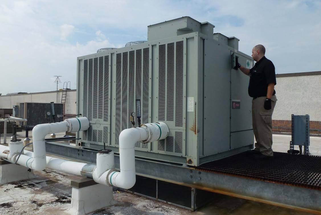 Technician inspecting large HVAC unit on a rooftop platform with white ductwork and pipes