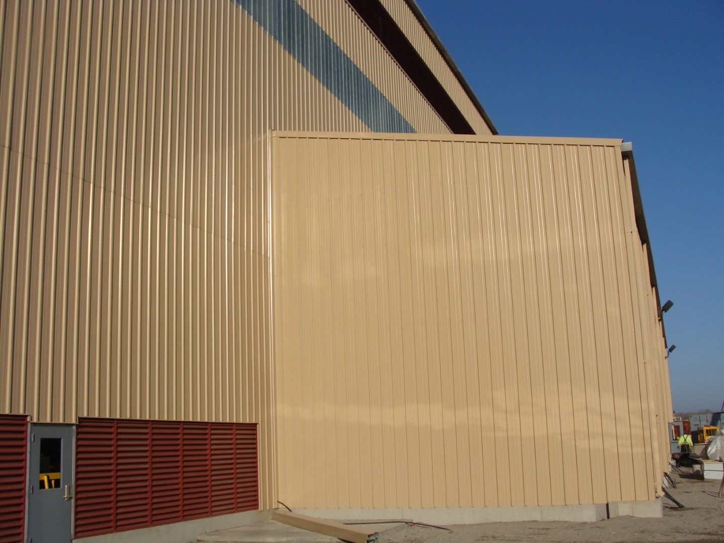 Tan corrugated industrial building with a large beige wall panel and red vented doors under blue sky