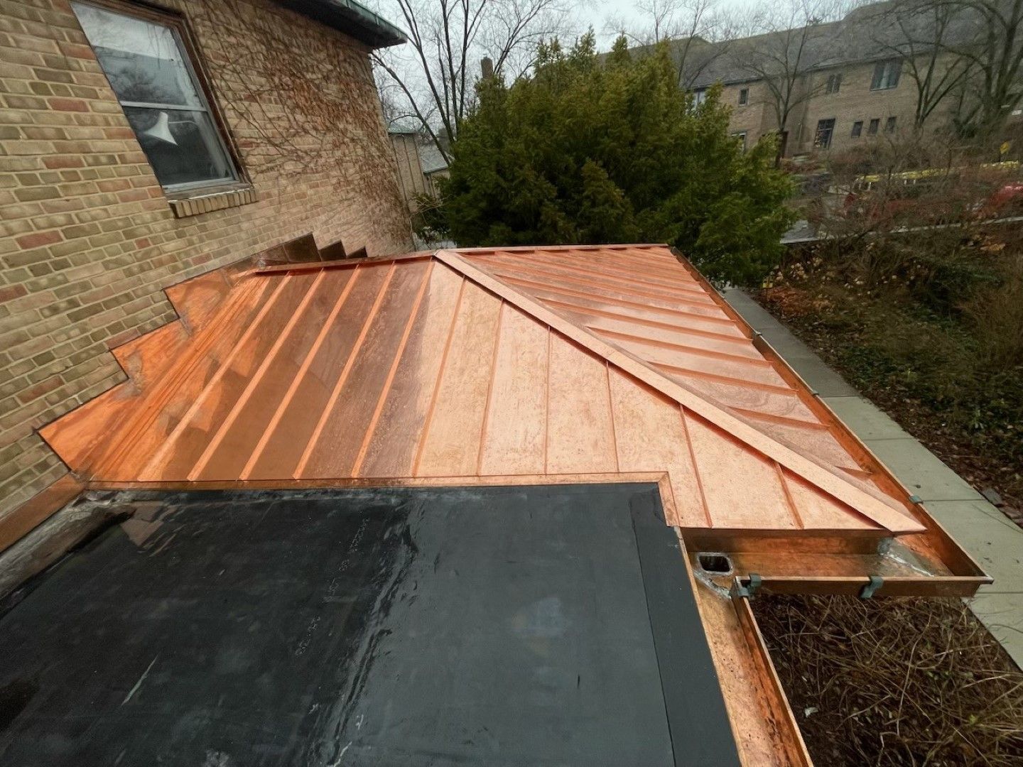 Copper standing-seam roof installed beside a brick house, with a black flat-roof section in the foreground