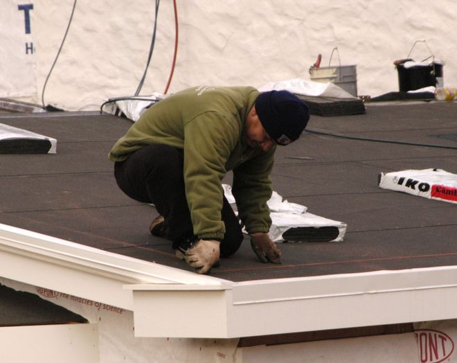 Worker in green jacket kneels on a rooftop, inspecting or repairing a white roof edge.