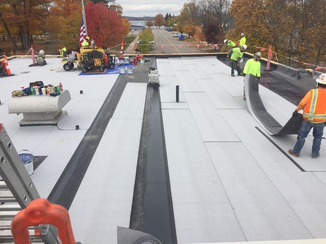 Workers installing roofing material on a flat roof with autumn trees in the background.