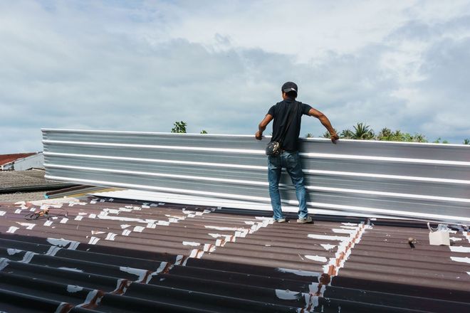 Worker installing metal roofing on a sloped roof under a cloudy sky