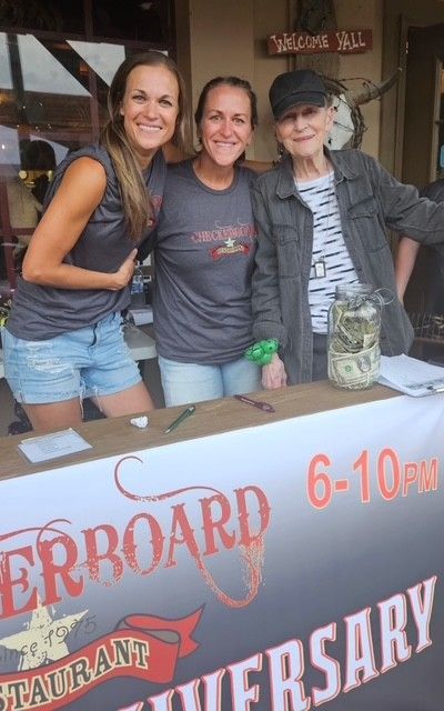 Three women smiling behind a table at the Outerboard Restaurant. The table has a jar for tips and an advertisement banner for the restaurant.