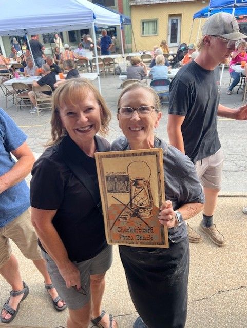 Two women smiling, holding a framed award at an outdoor event. People and tents are in the background.