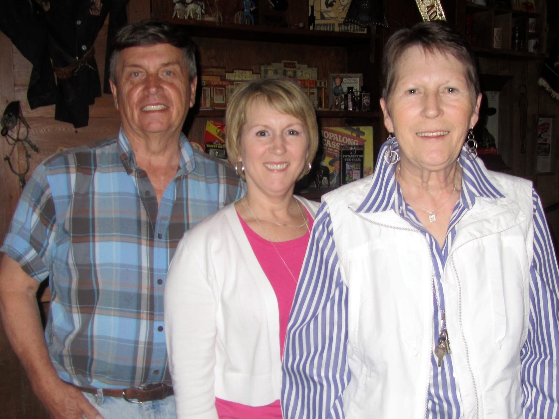 Three people smiling for a photo: a man in a plaid shirt, a woman in a pink top and white sweater, and a woman in a striped shirt and vest.
