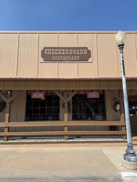 Checkerboard Restaurant exterior: brown building with a sign above the entrance, wooden porch, and a lamppost in front.