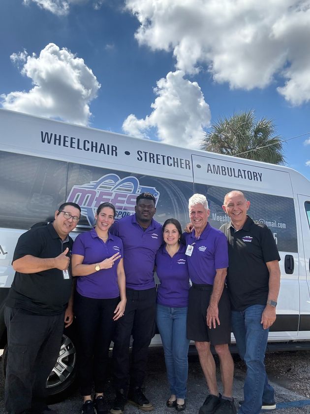 Six people stand in front of a medical transport van labeled Wheelchair, Stretcher, Ambulatory under a sunny sky.
