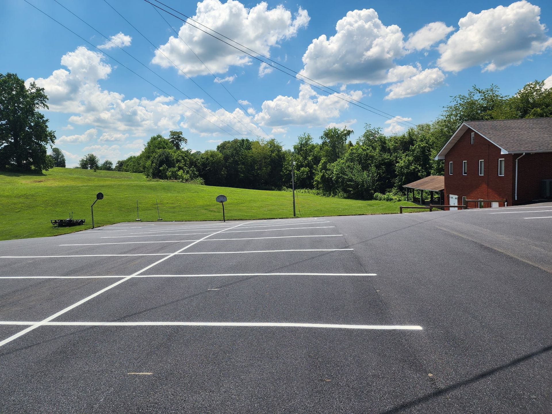 Empty asphalt parking lot next to a grassy field under a blue sky with puffy white clouds and trees.